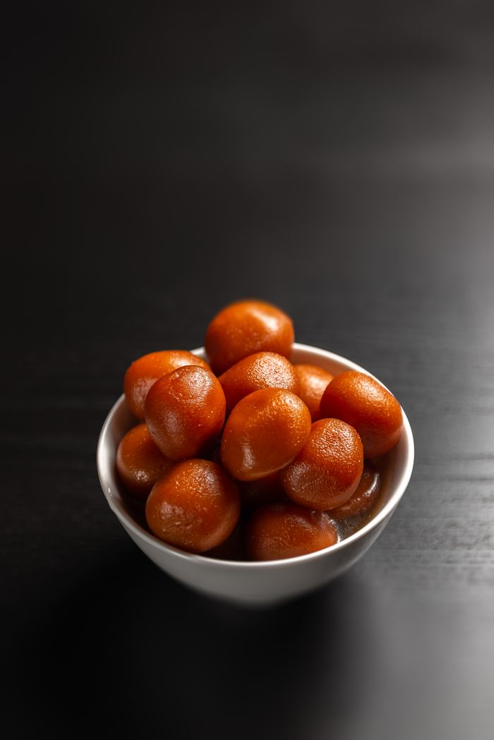 Close-up of traditional Indian sweet Gulab Jamun in a white bowl on black background.